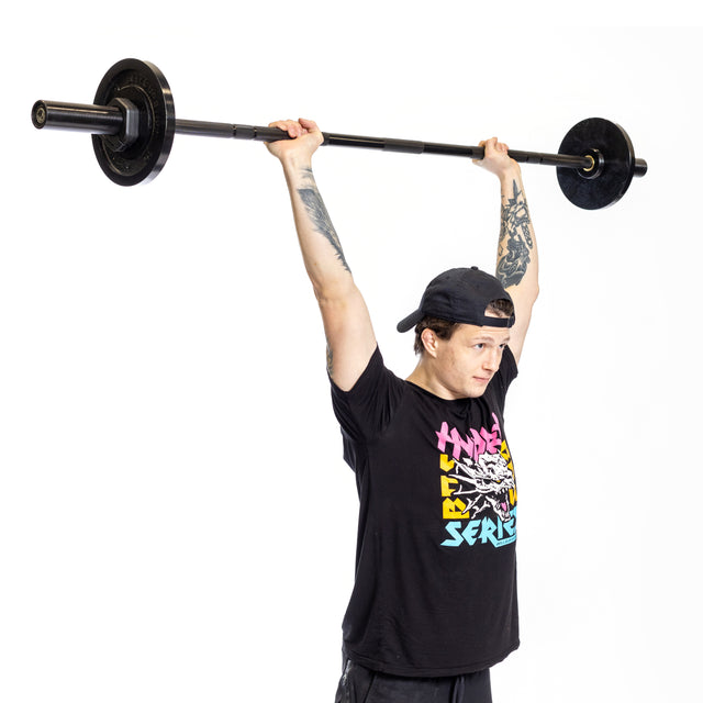 A tattooed individual in a black cap and graphic t-shirt lifts a Bells of Steel Short Rackable Barbell overhead against a white background, demonstrating strength. Ideal for compact home gyms, the bar supports up to 900 lbs.