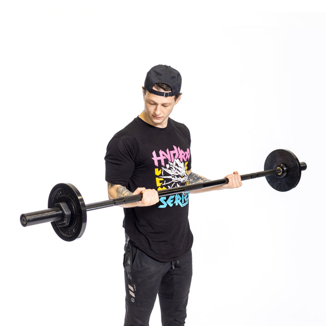 A person wearing a black cap, black t-shirt with vibrant text, and black pants lifts a Bells of Steel Short Rackable Barbell. Their focus is on the barbell, gripping it with both hands against a minimalist white backdrop resembling a home gym ideal for any power rack.