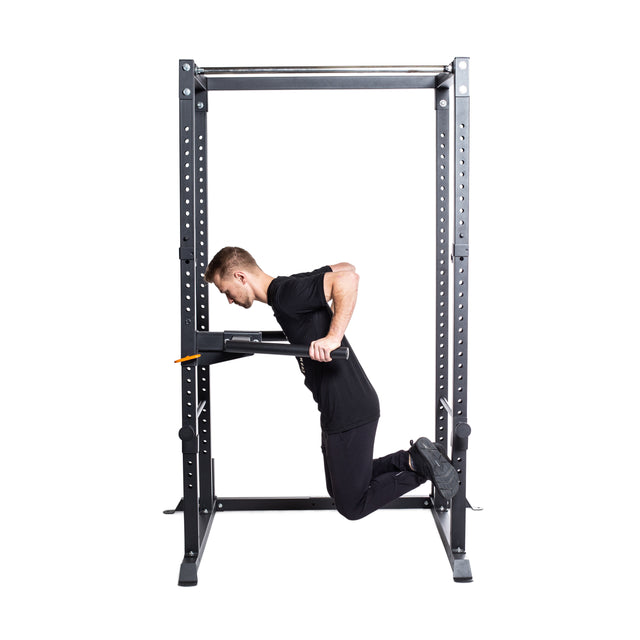 A man in black workout clothes does dips on parallel bars attached to a Bells of Steel Residential Power Rack (2.3" x 2.3", ⅝" HOLES) in his home gym, with his knees bent and feet crossed behind him.