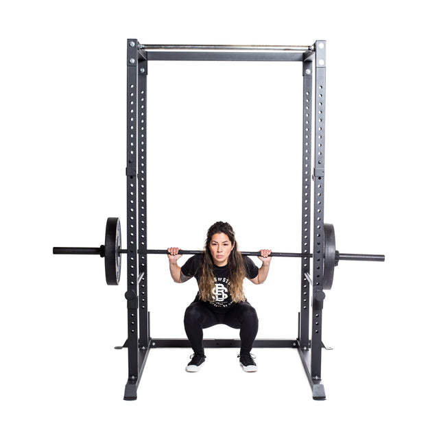 A woman with long hair performs a squat with a loaded barbell on her shoulders inside a Bells of Steel Residential Power Rack (2.3" x 2.3", ⅝" holes), the bar resting on j-cups, against a plain white background.