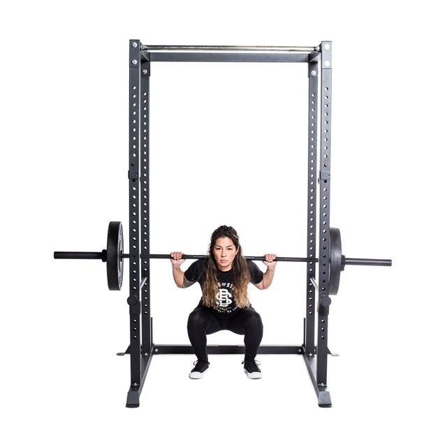 A woman with long hair performs a squat with a loaded barbell on her shoulders inside a Bells of Steel Residential Power Rack (2.3" x 2.3", ⅝" holes), the bar resting on j-cups, against a plain white background.