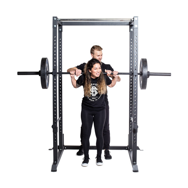 A woman prepares to squat with a barbell on her shoulders in the Bells of Steel Residential Power Rack (2.3" x 2.3", ⅝" holes), while a man spots her for safety; both wear black athletic clothes against a plain white background.