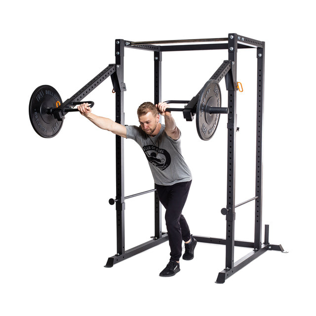A man in athletic wear uses a weightlifting machine with rotating arms and plates, set up inside the Bells of Steel Residential Power Rack (2.3" x 2.3", ⅝" holes)—a versatile addition to any home gym.