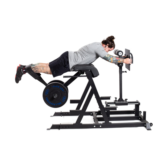 A tattooed individual with a bun works out on the Bells of Steel Reverse Hammer 2 In 1 GHD And Reverse Back Extension, targeting their posterior chain. Wearing a gray shirt, black shorts, and sneakers, they perform reverse back extensions with weights against a white backdrop.