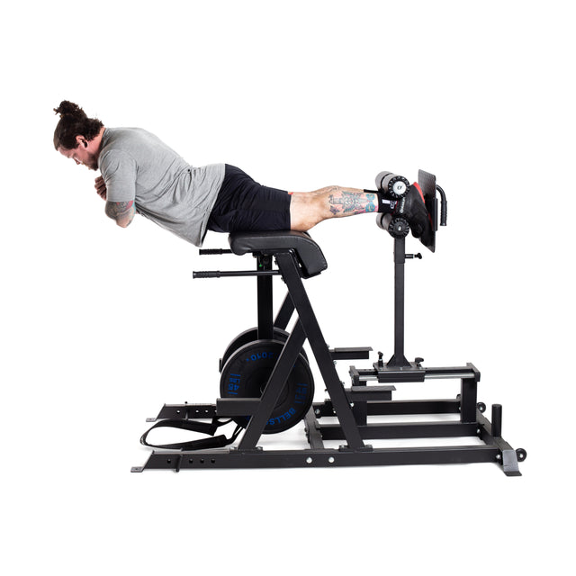 A person executes a reverse back extension on the Bells of Steel's Reverse Hammer 2 In 1 GHD and Reverse Back Extension. Wearing a gray shirt, black shorts, and socks, they use this robust machine with blue weights and a black frame to effectively target the posterior chain.