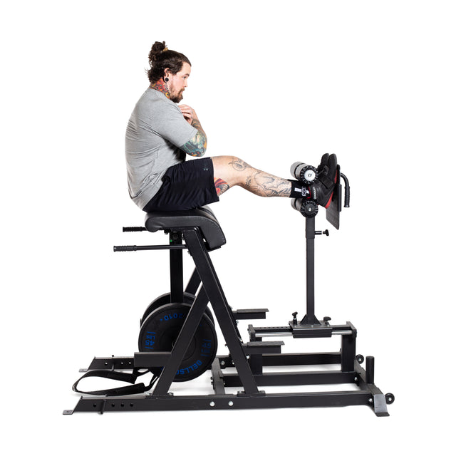 A tattooed individual with a ponytail, wearing a grey shirt and black shorts, is sitting on the Bells of Steel Reverse Hammer 2 In 1 GHD And Reverse Back Extension. Their arms are crossed and legs extended forward, performing an exercise for the posterior chain against a white background.
