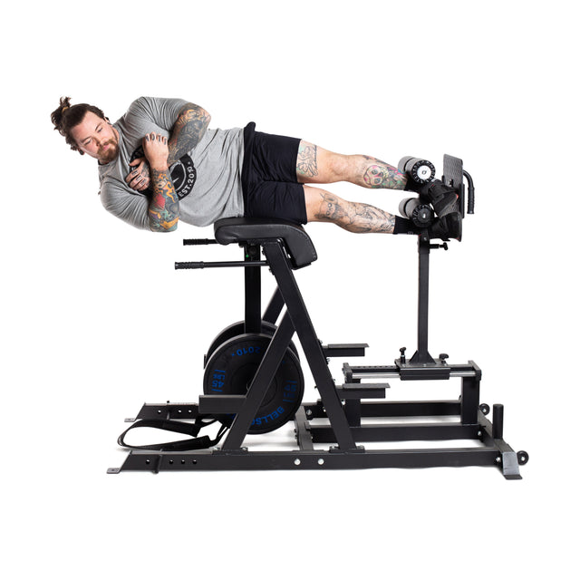 A tattooed individual with a bun works out on a Bells of Steel Reverse Hammer 2 In 1 GHD And Reverse Back Extension. Wearing a gray shirt and black shorts, they're positioned sideways, lying on the padded section with feet secured, focusing intently on strengthening their posterior chain.