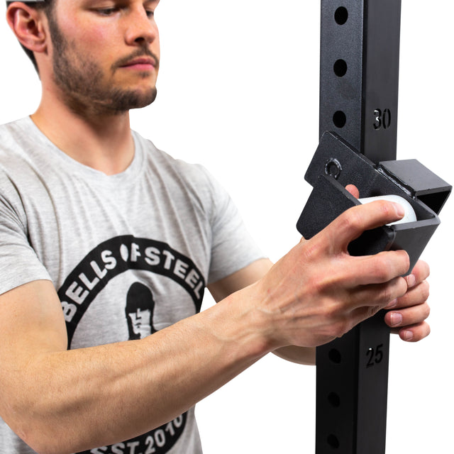 Wearing a Bells of Steel shirt, a man adjusts Roller J-Cups on a black numbered squat rack, demonstrating their durable nylon rollers for safe bench press and lifting sessions.