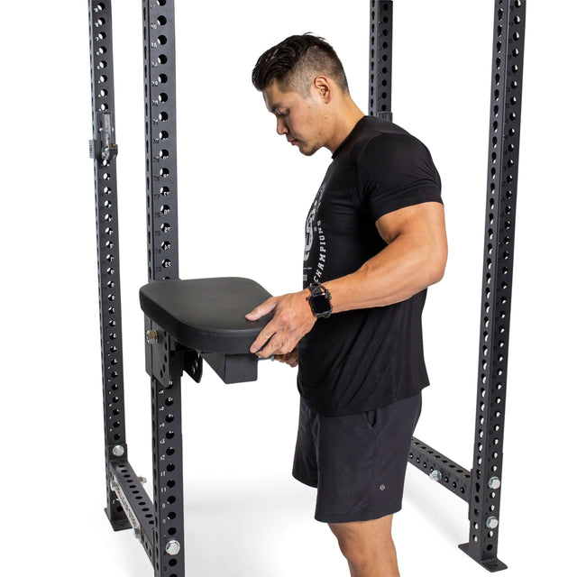 A man in black athletic wear adjusts a Bells of Steel Seal Row Pad Rack Attachment on a metal power rack, preparing the gym equipment for use.