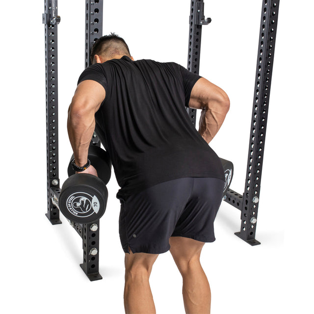 A person uses the Bells of Steel Seal Row Pad Rack Attachment inside a power rack to perform a bent-over row with two dumbbells, wearing a black shirt and shorts, facing away from the camera.