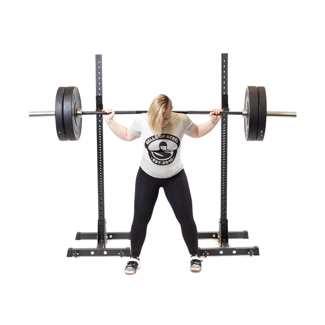A blond-haired person in a "House of Steel" t-shirt and black leggings prepares to squat with a loaded barbell on Bells of Steel Squat Stands (2.3" x 2.3", ⅝" holes), perfect for home gyms or weightlifting clubs, against a white background.