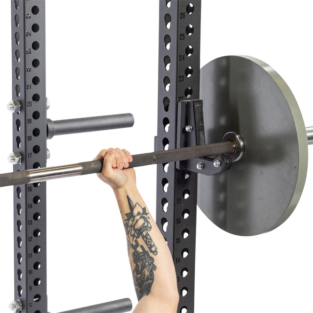 A person with a tattooed arm grips a barbell on a squat rack equipped with Bells of Steel Sandwich J-Cups, providing secure positioning and multiple adjustable height options.