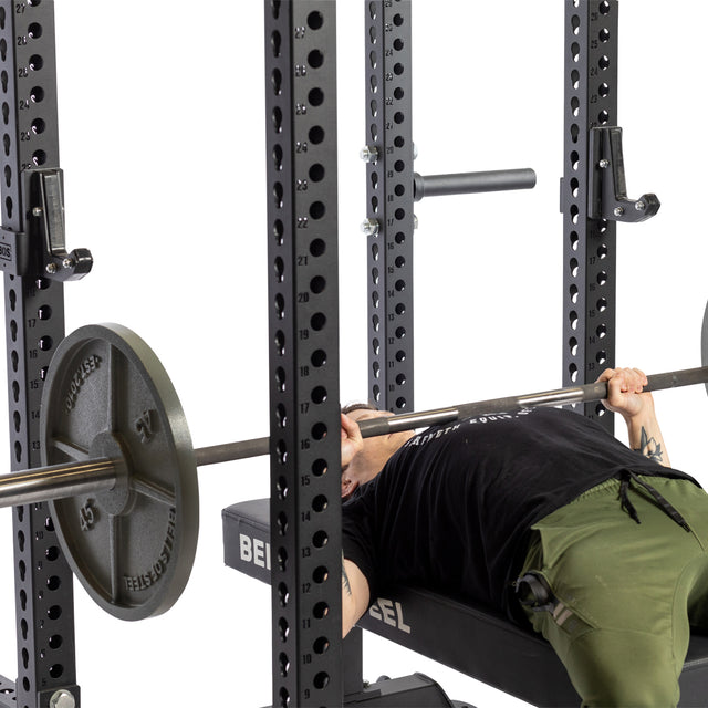 A person in a black shirt and green pants is lying on a bench inside a power rack, gripping a barbell resting on Bells of Steel Sandwich J-Cups, preparing for a bench press with heavy-duty rack attachments.
