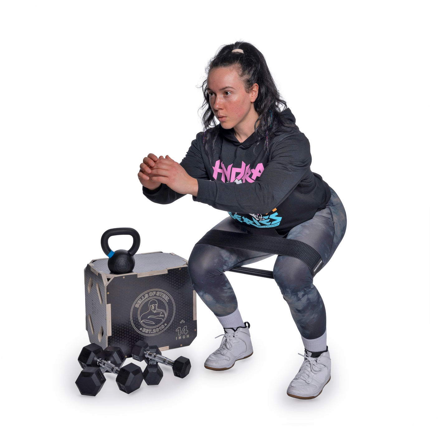 A woman in athletic wear performs a squat with arms forward near a box labeled "Bells of Steel Strength Circuit Starter Kits," with several dumbbells on the floor against a plain white background.