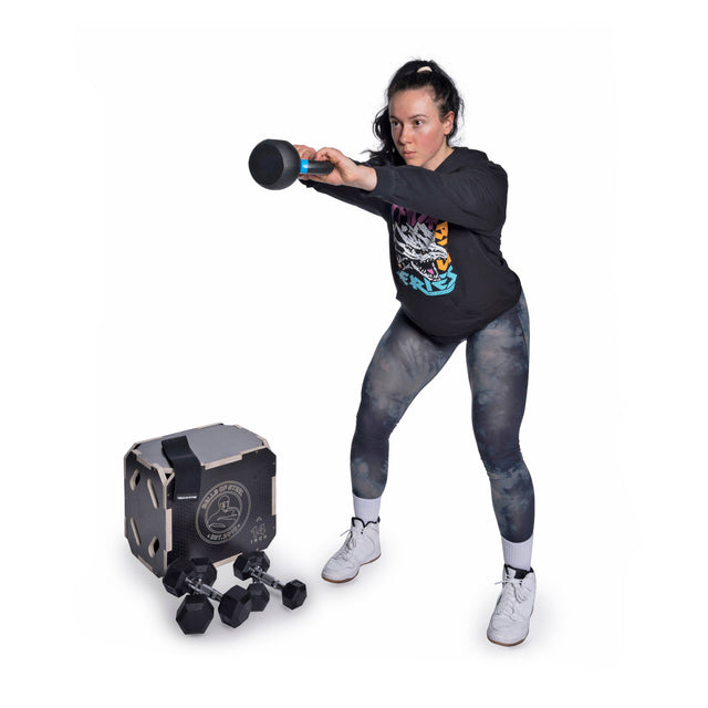 A woman in athletic wear swings a macebell with both hands beside a fitness cube and several dumbbells, all from the Bells of Steel Strength Circuit Starter Kits—perfect inspiration for your next home workout.