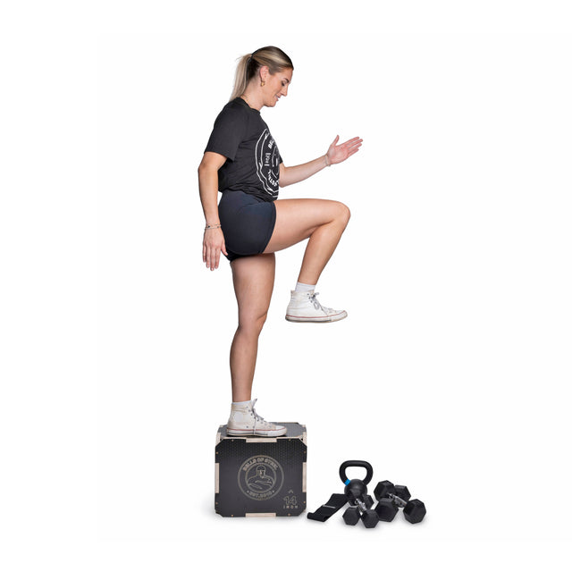 A woman in athletic wear demonstrates the Bells of Steel Strength Circuit Starter Kits, stepping onto a workout box with dumbbells and kettlebells beside her—ideal for home workouts—set against a white background.