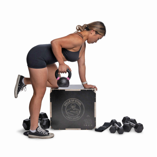 A woman in athletic wear uses a kettlebell while resting on a plyo box, with Bells of Steel Strength Circuit Starter Kits and dumbbells nearby—ideal for home workouts against a white backdrop.