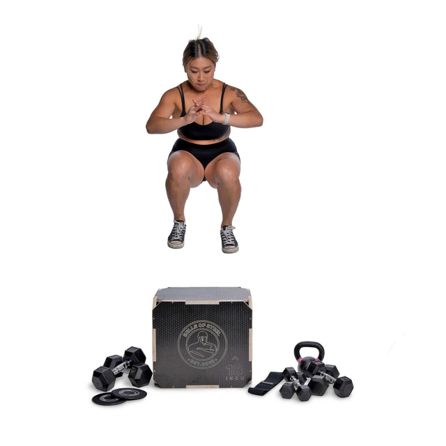 Wearing athletic gear, a woman performs a squat jump above a box surrounded by Bells of Steel Strength Circuit Starter Kits—featuring dumbbells, resistance bands, weight plates, kettlebell, sliders, and more—for an intense home workout.