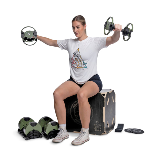 A woman works out at home, sitting on a plyometric box and lifting round, green dumbbells. She wears a graphic tee, shorts, and sneakers. Bells of Steel Strength Circuit Starter Kits and fitness gear are arranged nearby.