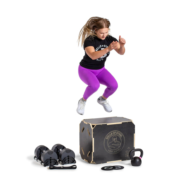 A woman in purple leggings and a black shirt jumps onto a plyo box. Bells of Steel Strength Circuit Starter Kits—dumbbells, a kettlebell, and weight plates—are on the floor before her, all set against a white background.
