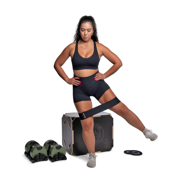 A woman in athletic wear uses a resistance band for a leg exercise, with Bells of Steel Strength Circuit Starter Kits, dumbbells, and a step platform nearby, creating an inspiring home workout scene on a white background.