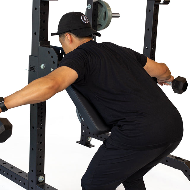 A person in black workout gear performs a dumbbell row on an incline bench inside a power rack, using the Bells of Steel Seal Row Pad Rack Attachment for chest support and added stability while holding dumbbells.