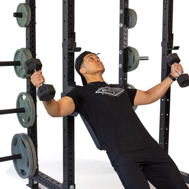 A man in black athletic wear performs an incline dumbbell bench press using the Bells of Steel Seal Row Pad Rack Attachment with weight plates loaded on a gym power rack.