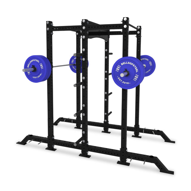 The Bells of Steel Hydra 2-Sided Rack is a black metal power rack with a barbell and blue weight plates at shoulder height, ideal for squats and bench presses in your modular home gym.
