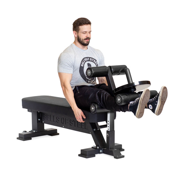 A man in a gray T-shirt uses the Bells of Steel Hero Heavy-Duty Weight Bench (1,000lb max capacity) with a leg extension attachment, lifting weighted pads while seated. The scene is set against a plain white background.