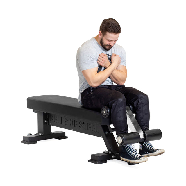 A man sits on the Bells of Steel Hero Heavy-Duty Weight Bench, arms crossed, ready for an ab exercise on the robust bench with a 1,000lb max capacity.