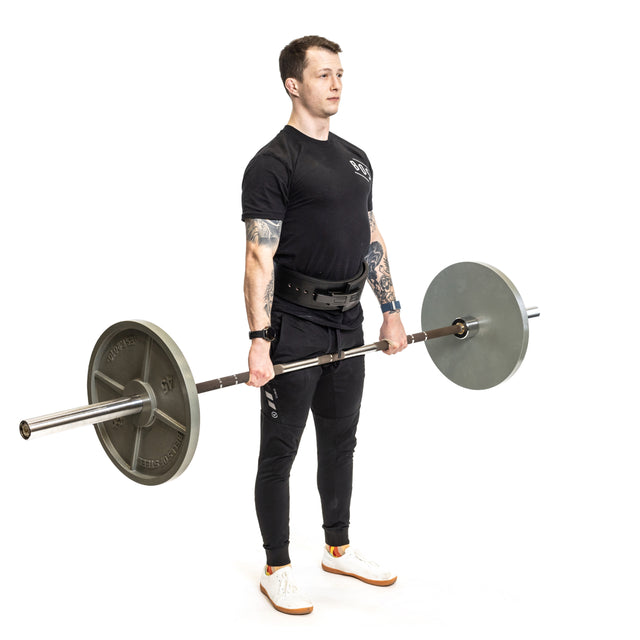 A man stands upright holding the Bells of Steel Hydra Seven Ring Bar, which features a 2000lb capacity, in front of his thighs. He wears a black t-shirt, black pants, white shoes, and a lifting belt against a white background.