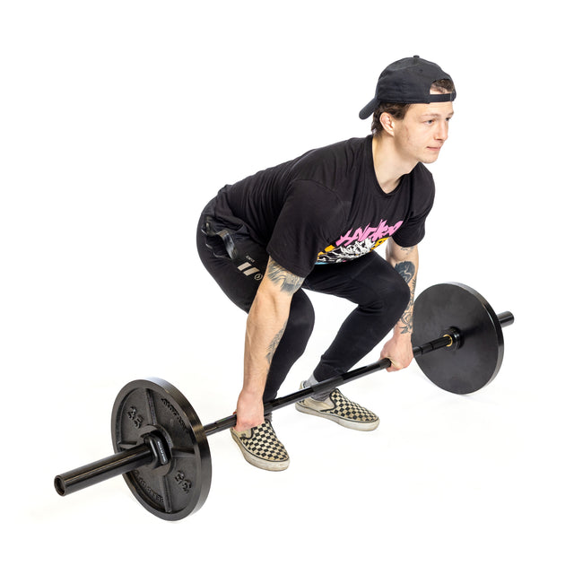 A young person in a black t-shirt, black pants, checkered slip-ons, and a backwards cap prepares to deadlift the Bells of Steel Short Utility Barbell in a home gym with a white background.