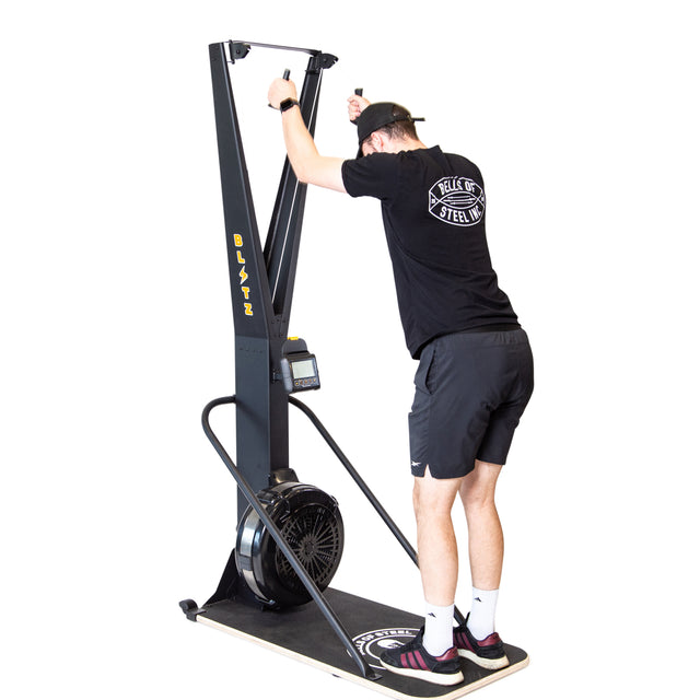 Wearing a black T-shirt, shorts, and cap, a person uses the Bells of Steel Blitz Ski Trainer, gripping the handles and leaning forward on the platform for a full-body workout.