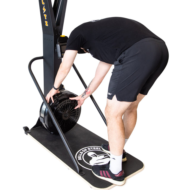 Wearing black athletic gear, a person adjusts the fan at the base of the Bells of Steel Blitz Ski Trainer, which sits on a mat displaying the brand's logo against a white background.