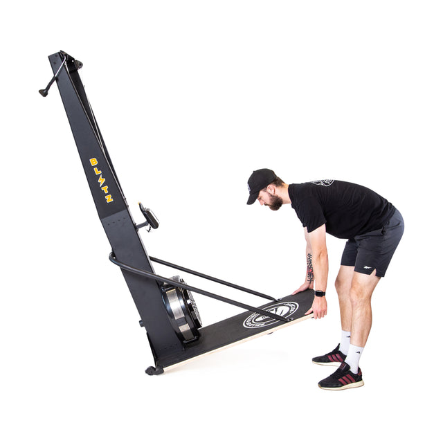 A man in athletic wear lifts the Blitz Ski Trainer by Bells of Steel, positioning it against a plain white background—perfect for simulating cross-country skiing and achieving a full-body workout.