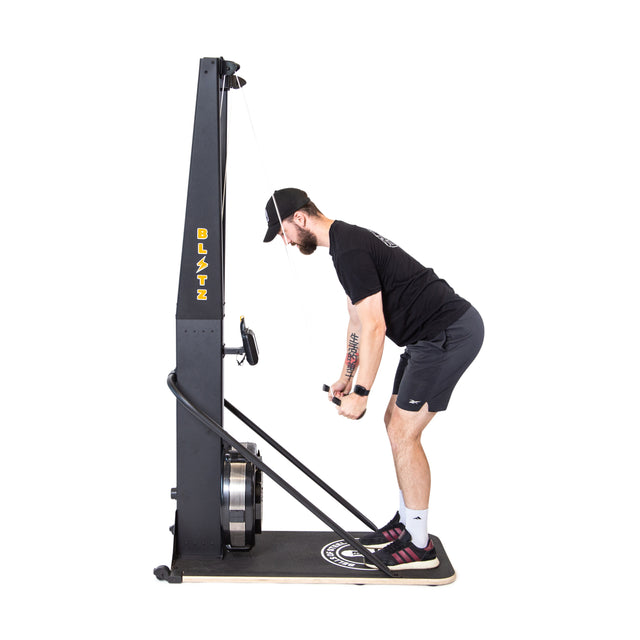 A man in athletic wear uses the Bells of Steel Blitz Ski Trainer, mimicking cross-country skiing as he powerfully pulls the handles downward while slightly bent forward, knees flexed for an intense full-body workout.