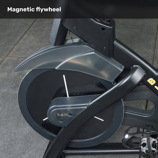 Close-up of the Bells of Steel Blitz Indoor Cycling Bike’s magnetic flywheel and pedal, showing its dark frame and gray cover. “Magnetic flywheel” is displayed in white on this adjustable exercise bike.