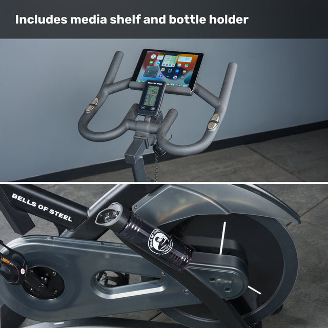 Close-up of the Bells of Steel Blitz Indoor Cycling Bike displaying its media shelf with tablet, water bottle holder, pedals, and flywheel, plus text above: "Includes media shelf and bottle holder.