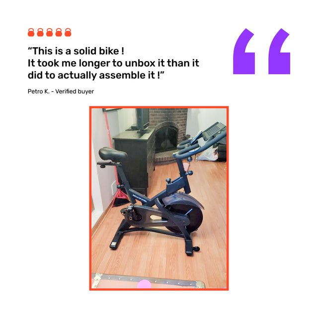 A Bells of Steel Blitz Indoor Cycling Bike stands on a wooden living room floor by a fireplace, with a review from verified buyer Petro K above, highlighting the bike’s easy assembly.