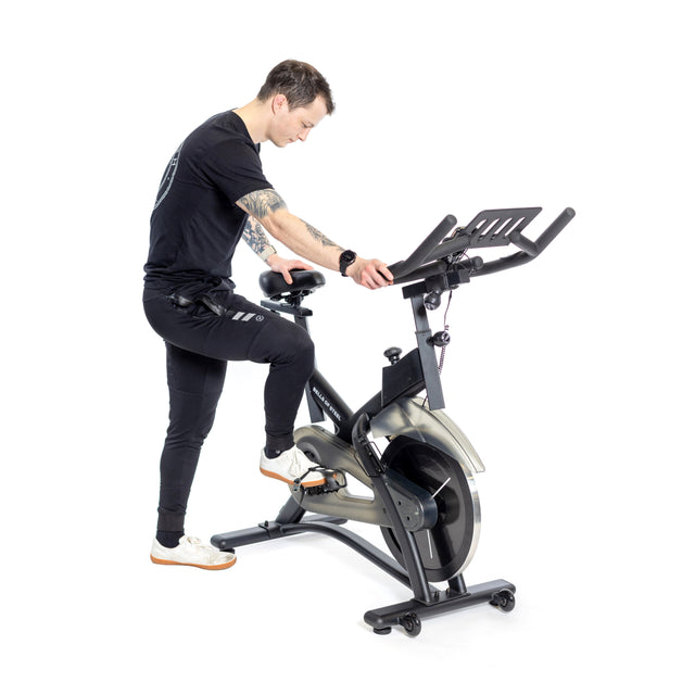 A tattooed, short-haired individual in black athletic wear adjusts the handlebars of a Bells of Steel Blitz Indoor Cycling Bike with magnetic resistance. The black and silver bike is striking against the plain white background.