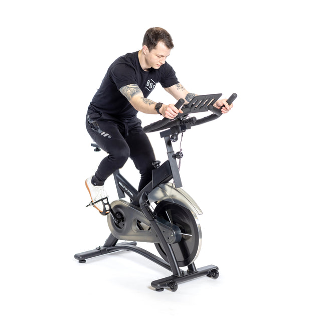 A person in black T-shirt and pants is riding a Bells of Steel Blitz Indoor Cycling Bike with magnetic resistance on a white background. They have short hair, appear focused, and grip the bike's adjustable handlebars while leaning slightly forward.