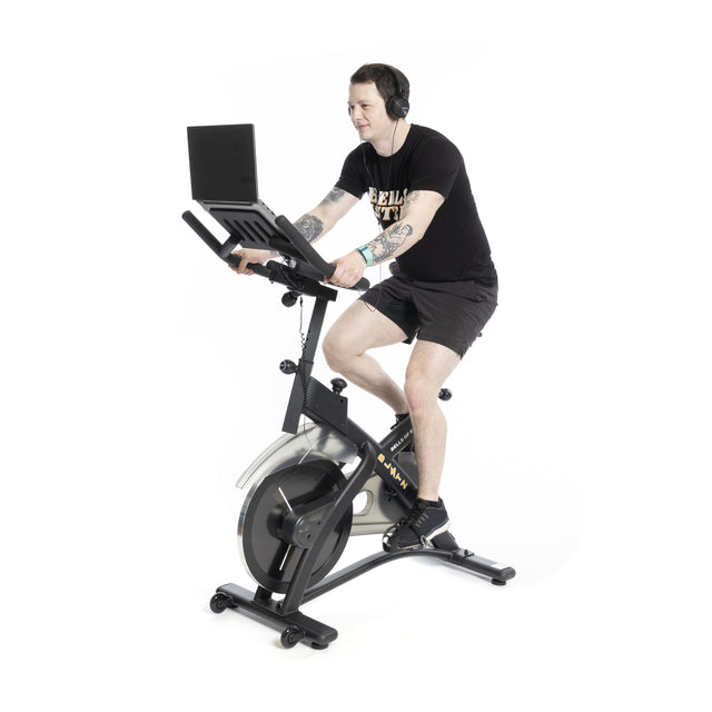 Dressed in a black T-shirt and shorts, a person exercises on the Bells of Steel Blitz Indoor Cycling Bike with adjustable handlebars and magnetic resistance, while watching a laptop, all set against a white background.