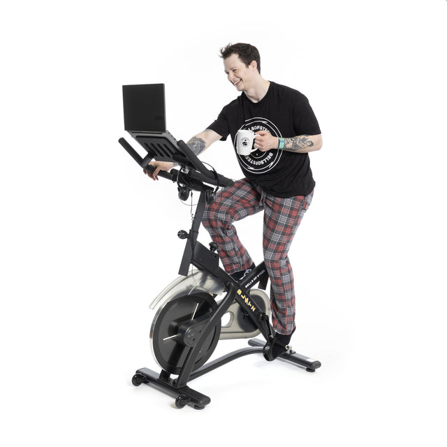 A person pedals a Bells of Steel Blitz Indoor Cycling Bike, coffee mug in hand and typing on a laptop. Its adjustable handlebars and magnetic resistance enable easy multitasking. Clad in a black T-shirt and plaid pajama pants, they cycle against a plain white backdrop.