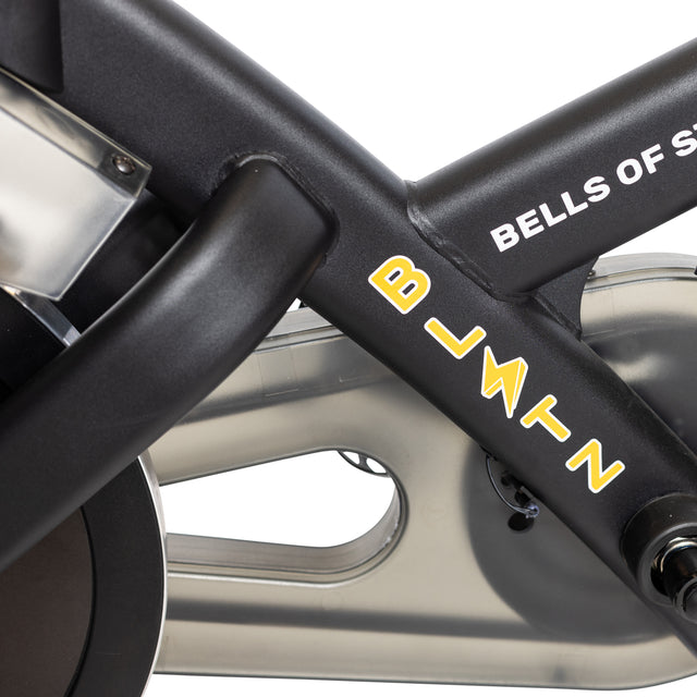 Close-up of the Blitz Indoor Cycling Bike's black frame with "Bells of Steel" in white, a yellow and white logo, partially visible pedals, adjustable handlebars, and a metal component in the background.