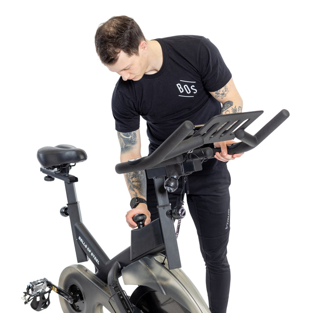A tattooed person in a black Bells of Steel t-shirt and pants adjusts the handlebars of a Blitz Indoor Cycling Bike, focused on achieving the perfect setup against a plain white background.