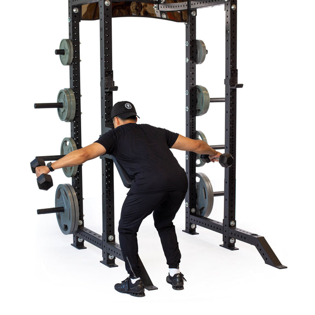 Wearing black workout clothes, a person does a bent-over dumbbell raise in front of a squat rack with the Bells of Steel Seal Row Pad Rack Attachment and weight plates.