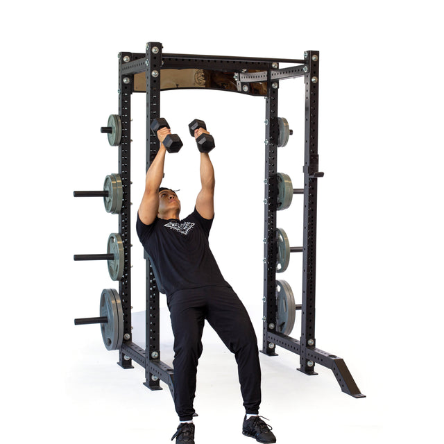 A person in black workout clothes performs an incline dumbbell press while leaning against a squat rack fitted with the Bells of Steel Seal Row Pad Rack Attachment, set against a white background.