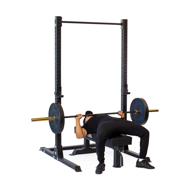 A person lies on a bench under the Bells of Steel Hydra Squat Stand - Prebuilt (3" x 3", ⅝" Holes), performing a barbell bench press with weights; the plain white background highlights this essential home gym equipment.