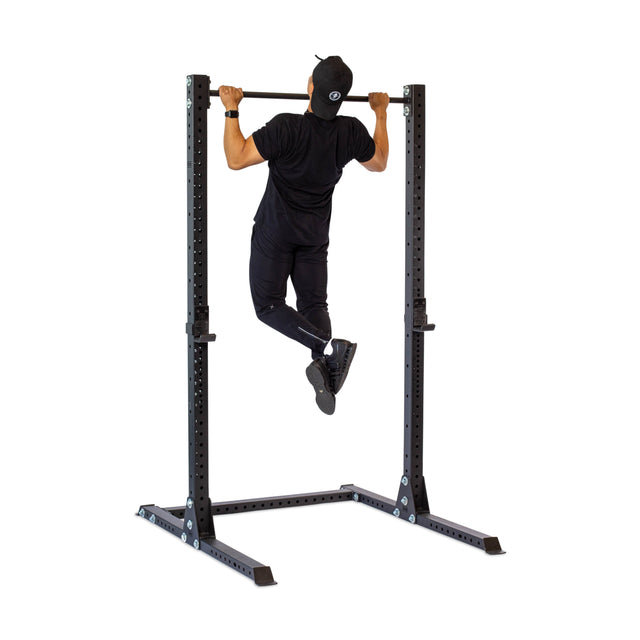 A person in black athletic wear and a cap performs a pull-up on the Bells of Steel Hydra Squat Stand - Prebuilt (3" x 3", ⅝" Holes); the sturdy metal frame resembles racks found in home gyms. The image is taken from behind.