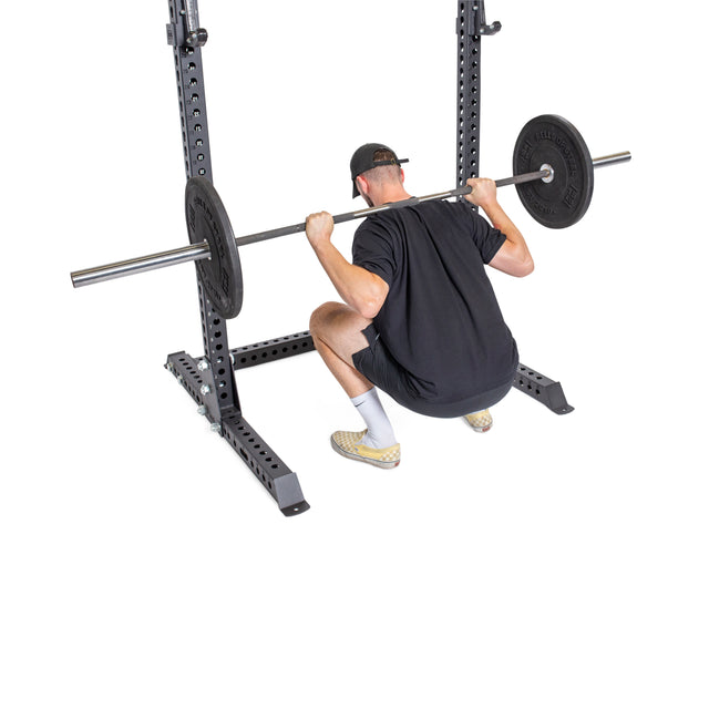 A person in a black shirt, shorts, and cap performs a deep squat with a barbell on their shoulders using the Bells of Steel Manticore Squat Stand Builder (3" x 3", 1" Holes) against a white background, showcasing a sleek home gym setup.